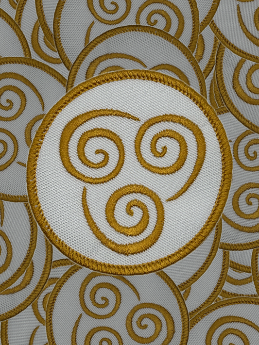 Gold embroidered triskele symbol on a white fabric background with gold swirling patterns