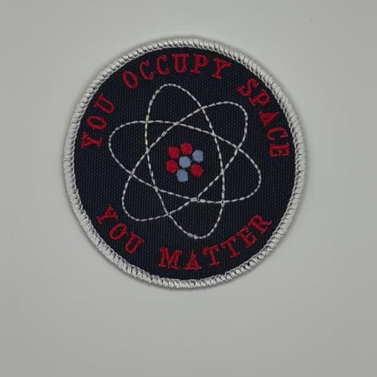 Round patch with atomic symbol and text 'YOU OCCUPY SPACE YOU MATTER' on a black background